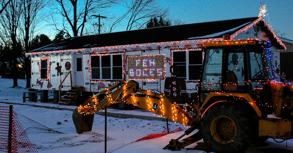 A partially constructed house is lit up with holiday lights with a decorated piece of heavy equipment in front of it