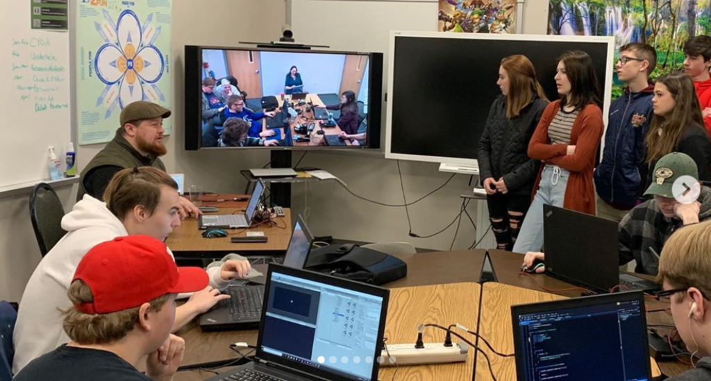 Visiting students stand in a classroom talking with the sitting teacher and students at laptops. A videoconferencing screen shows remote students and their teacher.
