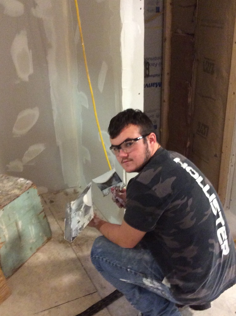 A student wearing safety glasses and holding plastering equipment looks at the camera.